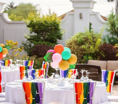 Mese festive cu scaune decorate în curcubeu și baloane colorate.