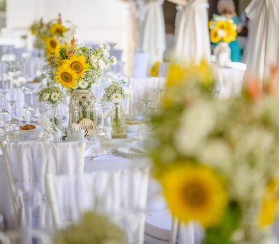 Mese aranjate cu flori și eticheta "Masa 2". Pahare și farfurii curate pe mesele acoperite cu alb.