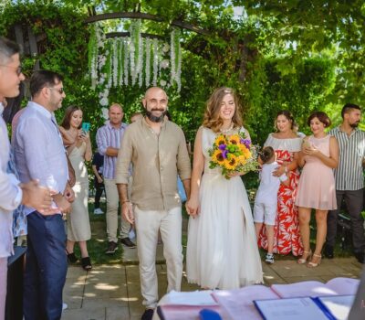 Cuplu căsătorit, ținându-se de mână, mergând pe culoar spre ceremonia de nuntă în aer liber, cu invitați și verdeață luxuriantă.