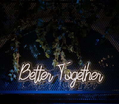 Un semn de neon alb spune "Better Together" pe un fundal întunecat, metalic, cu frunze verzi.
