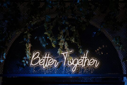 Un semn de neon alb spune "Better Together" pe un fundal întunecat, metalic, cu frunze verzi.