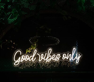Neon alb cu textul „Good Vibes Only” pe fundal negru cu vegetație.
