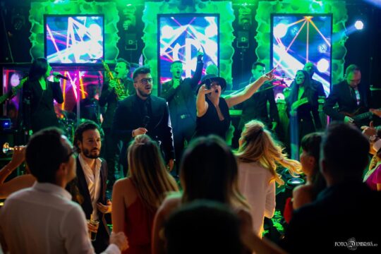 O trupă live interpretează în fața unei mulțimi la un eveniment, lumini verzi strălucind în fundal.