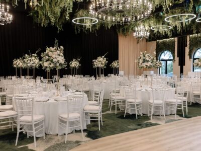 Sala de bal amenajată cu mese rotunde albe, scaune și aranjamente florale. Candelabre și plante decorative atârnă.