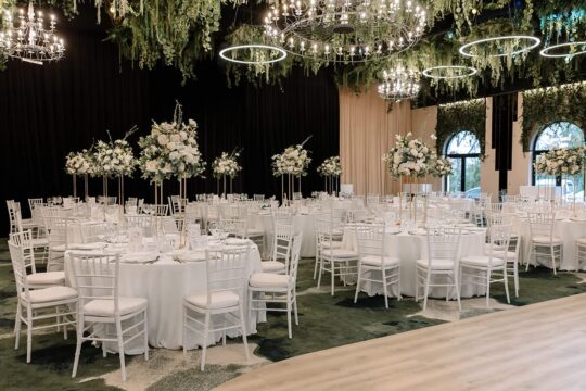 Sala de bal amenajată cu mese rotunde albe, scaune și aranjamente florale. Candelabre și plante decorative atârnă.