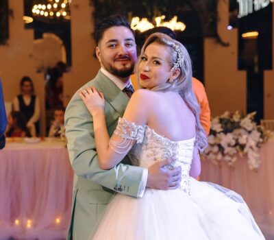 Un cuplu îmbrățișat, el cu costum verde pal, ea cu rochie albă la eveniment.