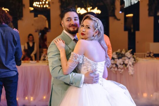 Un cuplu îmbrățișat, el cu costum verde pal, ea cu rochie albă la eveniment.