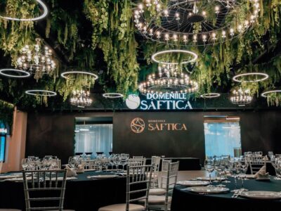 Interiorul "Domeniile Saftica" cu candelabre, vegetație, mese negre și sticlărie.