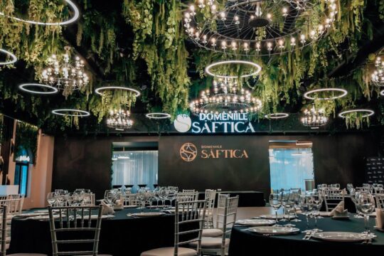 Interiorul "Domeniile Saftica" cu candelabre, vegetație, mese negre și sticlărie.
