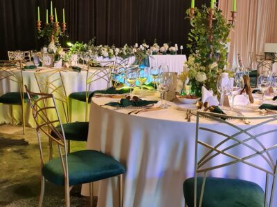 Mese rotunde cu fețe de masă albe, scaune aurii cu tapițerie verde, lumânări, aranjamente florale.