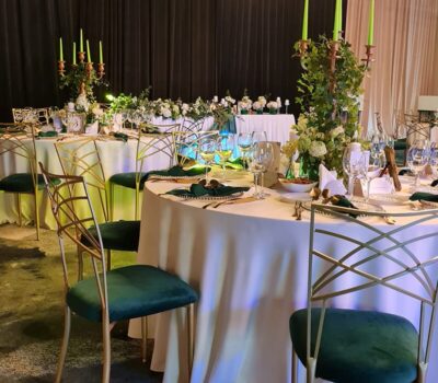 Mese rotunde cu fețe de masă albe, scaune aurii cu tapițerie verde, lumânări, aranjamente florale.