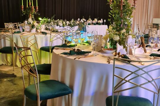 Mese rotunde cu fețe de masă albe, scaune aurii cu tapițerie verde, lumânări, aranjamente florale.