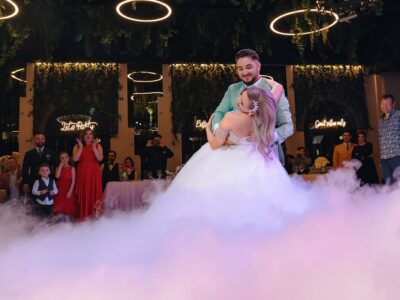 Cuplu la nunta, dansând pe o podea cețoasă. Pe fundal, oaspeți și lumini decorative.