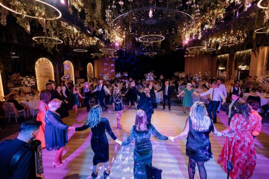 Într-o sală de bal împodobită, o adunare bucuroasă dansează, ținându-se de mână într-un cerc. Muzicienii cântă.