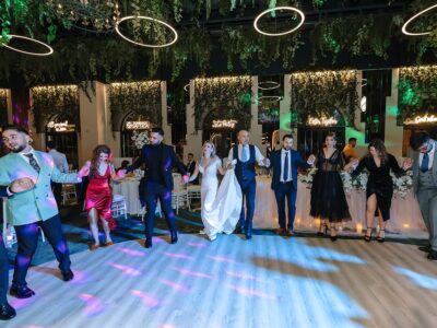 Un grup de oameni ținându-se de mână și dansând la o nuntă, în interior, cu instalații verzi și lumini circulare.