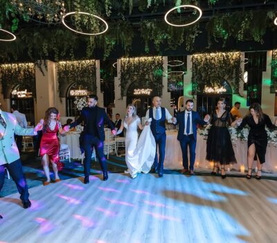 Un grup de oameni ținându-se de mână și dansând la o nuntă, în interior, cu instalații verzi și lumini circulare.