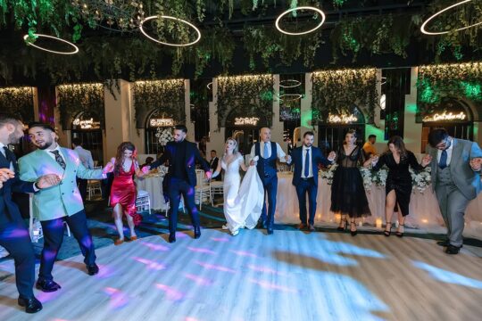 Un grup de oameni ținându-se de mână și dansând la o nuntă, în interior, cu instalații verzi și lumini circulare.