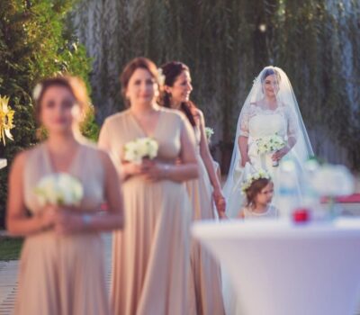 O mireasă cu un voal lung și domnișoare de onoare în rochii nude dețin buchete albe.