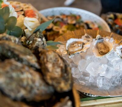 Străchini cu stridii, unele pe gheață, altele închise. Fundal cu sushi și decorațiuni florale.