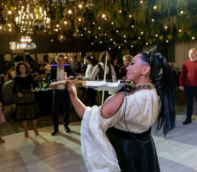 Femeie cântând la o vioară albă la o petrecere, oameni și lumini în fundal.