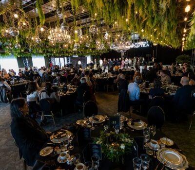 Sală de evenimente, cu mese aranjate, mulți oaspeți așezați și un grup de persoane pe scenă.