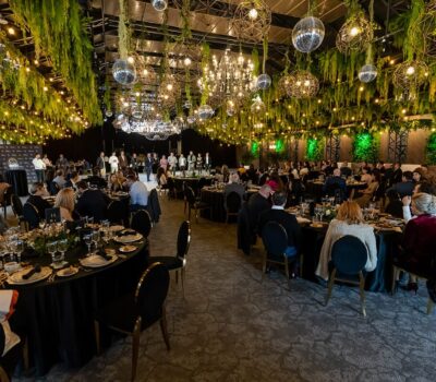 Interiorul unei săli de evenimente cu oameni așezați la mese rotunde. Plafonul e decorat cu verdeață și lumini.