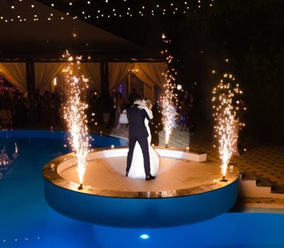 O mireasă și un mire dansează pe o platformă pe o piscină înconjurată de artificii și luminițe.