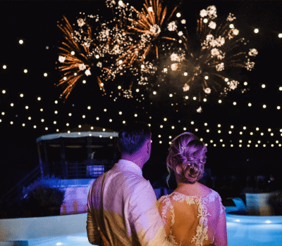 Un cuplu urmărește focuri de artificii deasupra unei piscine luminate. Lumini șirag atârnă sus.