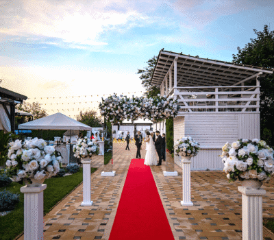 Nuntă în aer liber cu covor roșu, flori albe și cuplu căsătorit, lângă invitații la nuntă.