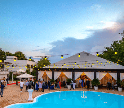 O petrecere sau nuntă în aer liber cu lumini, o piscină și o mulțime de oaspeți.