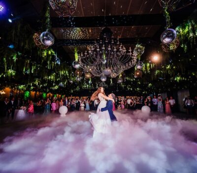 Un cuplu proaspăt căsătorit dansează pe un nor de gheață uscată, sub un candelabru într-o sală de bal, cu oaspeți în fundal.