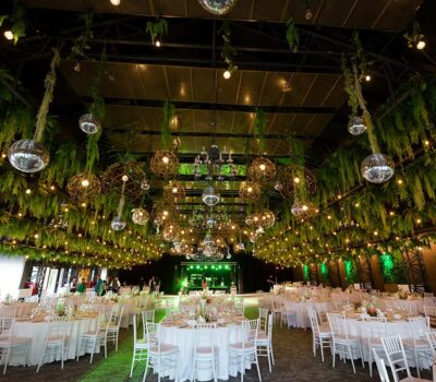 Interiorul decorat pentru eveniment cu mese rotunde, plante agățate, globuri disco și lumini. O scenă este situată în spate.