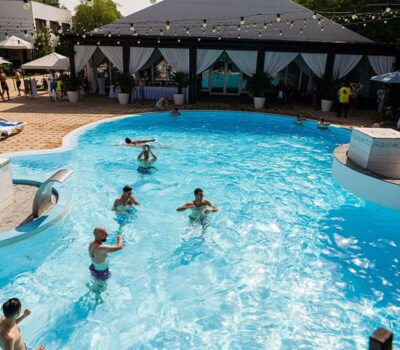 Oameni care se bucură de o piscină mare în aer liber, cu un pavilion în fundal.