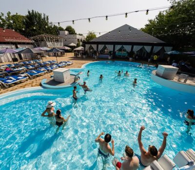 Oamenii se bucură de o piscină mare și plină de soare, cu șezlonguri și lumini suspendate.