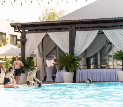 Oameni se relaxează lângă piscină, cu corturi și vegetație în fundal.