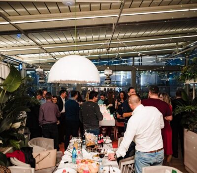 Un grup de oameni se adună în jurul unei mese lungi, într-un spațiu luminos și modern, pentru un eveniment social.