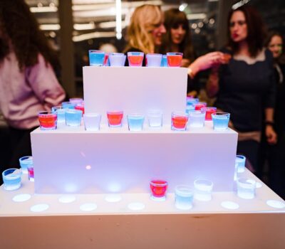 O piramidă albă cu shot-uri roșii și albastre, lumini sub și oameni în fundal.