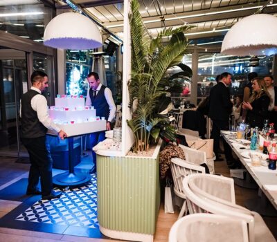 Doi ospătari cară o piramidă de shot-uri într-un restaurant aglomerat.