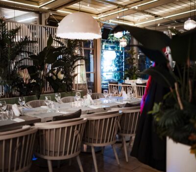 Interiorul unui restaurant elegant, cu mese aranjate și plante verzi.