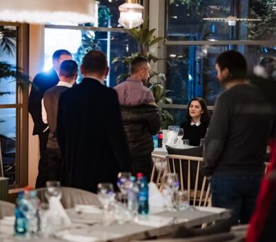 Într-o cameră, mai multe persoane stau lângă ferestre și mese aranjate.