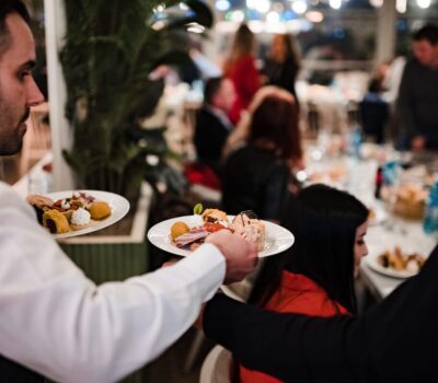 Un chelner poartă două farfurii cu aperitive la o adunare, cu oaspeți în fundal.