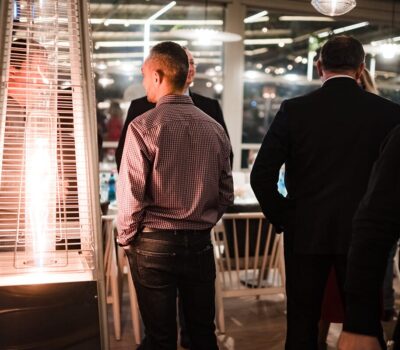 Persoane în interior, lângă un încălzitor terasă, purtând cămăși și costume întunecate, cu luminile slabe în fundal.