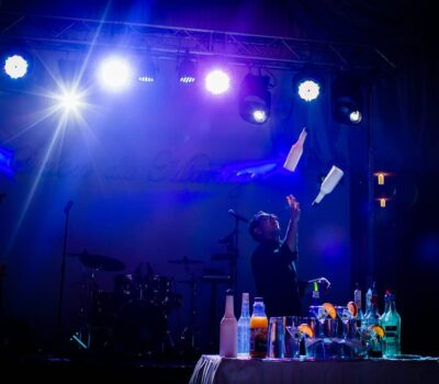 Un barman jonglează cu sticle pe o scenă iluminată cu lumini albastre și are o masă cu băuturi.