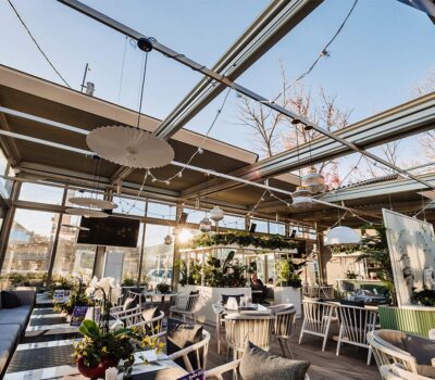 Interiorul unui restaurant luminos, cu ferestre mari, mese, scaune, plante și canapele.