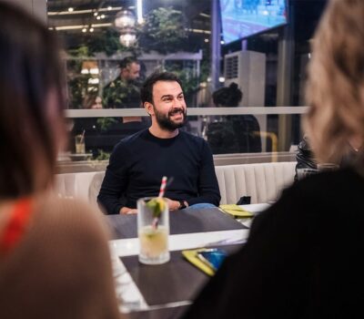 Un bărbat cu barbă zâmbește, stând la o masă între doi oameni. Un pahar cu băutură stă pe masă.