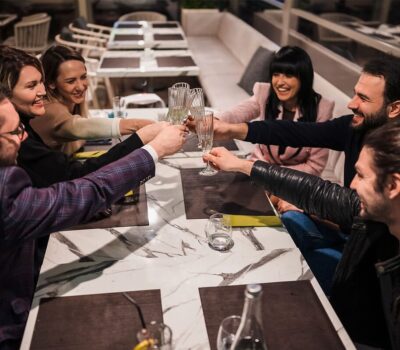 Șase prieteni ciocnesc pahare pe o masă de marmură într-un restaurant, zâmbind.