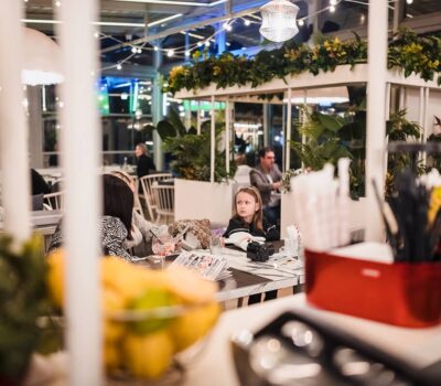 Interiorul unui restaurant. O fată stă la o masă cu adulți. Plantele sunt vizibile în fundal.