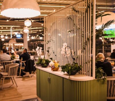 Interiorul unui restaurant cu persoane așezate la mese. Plante și corpuri de iluminat decorative.