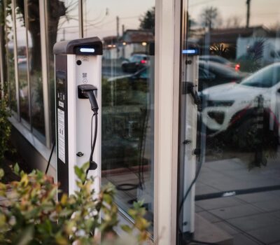 Stație albă de încărcare pentru vehicule electrice cu conector negru, reflectând vehicule în geamuri.