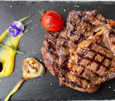 Friptură de vită aromată cu usturoi copt, roșii coapte, piure galben și flori comestibile pe o farfurie neagră.